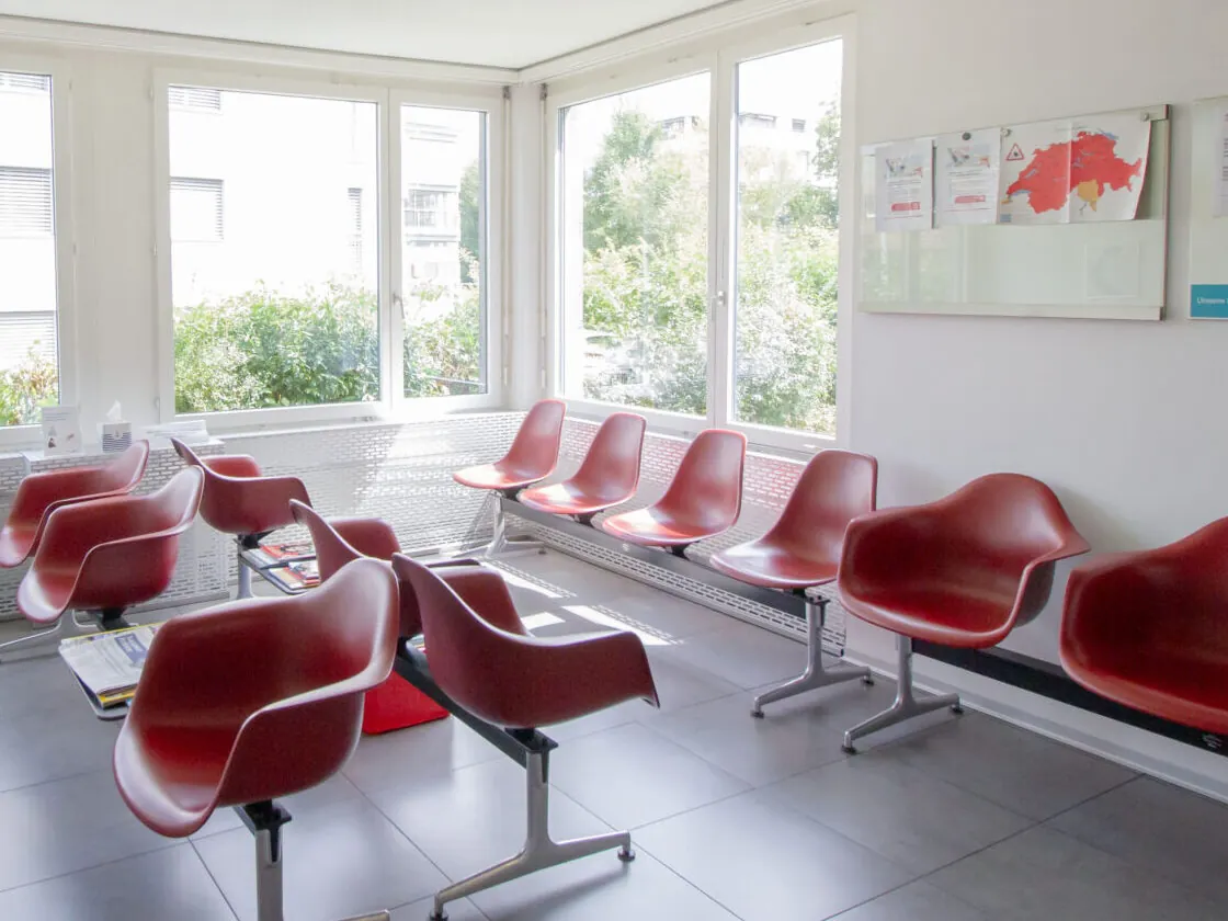Helles Wartezimmer der Hausarztpraxis Suhr mit roten Stühlen und Fensterfront